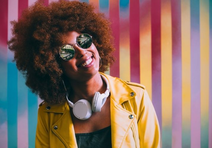 Uma jovem negra com óculos de sol espelhado, fone de ouvido no pescoço e uma jaqueta de couro amarela. Ela está sorridente na frente de uma parede listrada de amarelo, vermelho e azul, dando uma sensação de alegria.